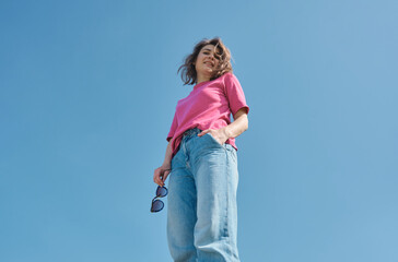 Beautiful carefree young woman with curly hair in pink t-shirt smiling and looking down at camera on blue sky background low angle shot.