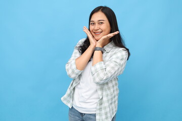 Fototapeta premium Portrait of a happy and smiling young Asian woman looking at the camera in a beautiful and flirtatious pose putting both hands under her chin, feeling amazed, standing isolated on a blue background.