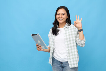 Young Asian woman holding computer digital tablet while gesturing ok sign with finger, smiling looking at camera, standing isolated on blue background.