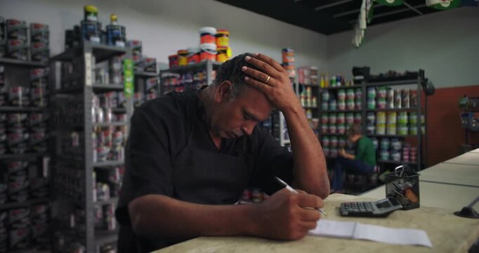 Male shop owner holding head in frustration while reviewing notes, deeply worried about financial challenges, small business struggles in retail store, pensive thoughtful expression