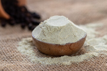 A wooden bowl filled with fine white powder, possibly flour or spice, on a burlap surface with scattered powder and blurred peppercorns in the background