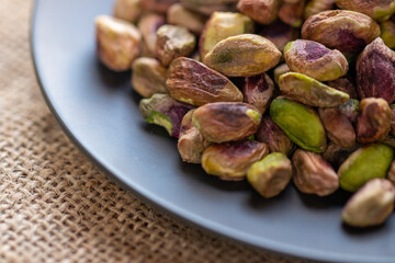 Closeup of a dark plate filled with roasted pistachios, showcasing their vibrant green and purple hues on a textured burlap background