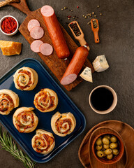 Freshly baked savory rolls with sliced sausage, blue cheese, hard cheese, peppercorns, olives and cup of red wine on serving board, festive gourmet snack, cozy evening kitchen appetizer in daylight