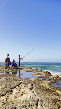 Men fishing on jetty of rugged coastline under clear blue sky
