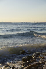 Sailboats on the horizon with waves crashing on a rocky shore at sunset
