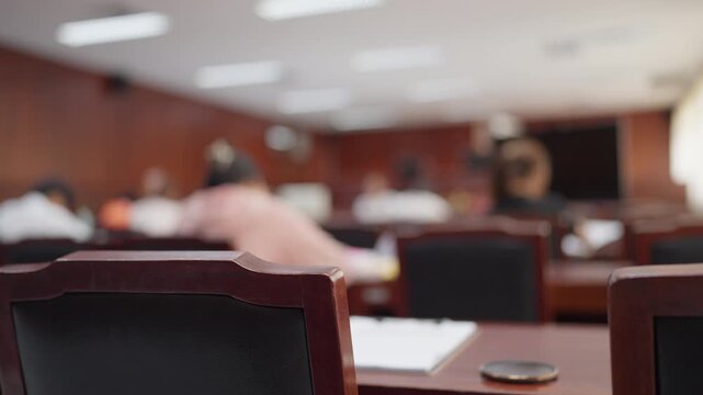 Wooden courtroom chair in focus with blurred people attending a legal trial, symbolizing law and justice.