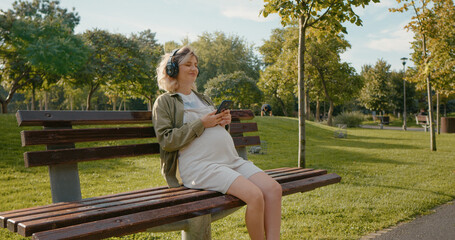 Pregnant with eyes closed is listening to music on bench in the park. Future mom enjoying nature while hearing songs in headphones.