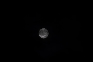 A close-up of the bright full Moon against the deep black night sky, with some clouds. The lunar disk is sharp and detailed, with the texture of the craters and lunar maria clearly visible.