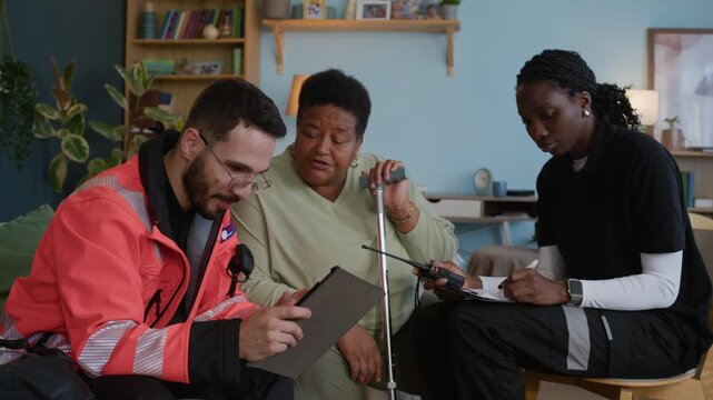 Medium long shot of male paramedic in orange uniform using digital tablet while talking to aged woman while female colleague filling medical form and radioing