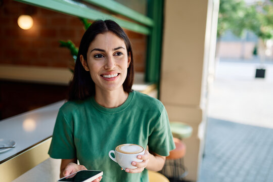 Portrait of a young woman girl sitting in cafeteria or coffee shop drinking coffee or cappuccino and using a smartphone mobile phone in the city and having fun - Powered by Adobe