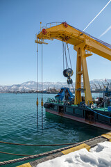 Fototapeta premium Yellow port crane lifting blue cargo on barge with mountain landscape backdrop