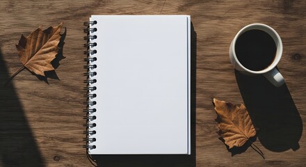 Overhead view of a blank spiral notebook, a warm cup of coffee, and autumnal leaves arranged on a rustic wooden table, perfect for creative work or planning sessions