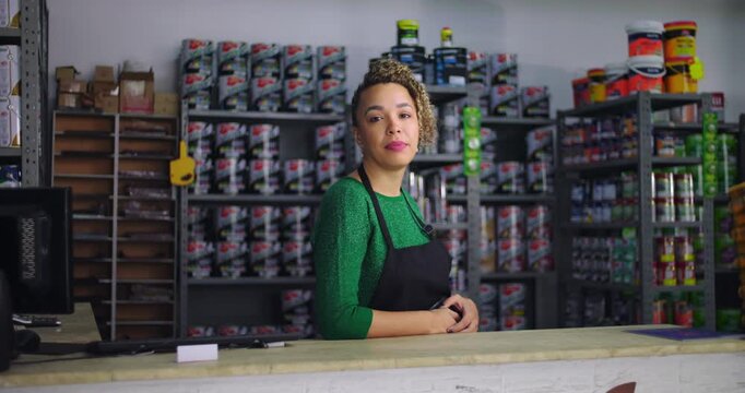 Confident woman standing at counter in local small business hardware store, surrounded by neatly arranged shelves of paint and tools, conveying professionalism and focus