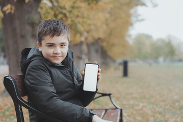 Smiling boy holding smartphone with blank white screen in autumn park, perfect for mobile app mockup, digital education concept, children technology usage, and seasonal lifestyle content.