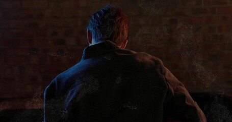Standing man wearing dark canvas jacket against brick wall in studio, with colored accent lighting