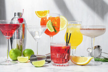 Glasses of tasty cocktails, ingredients, shaker, sieve and jigger on white marble table