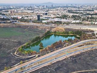 Drone photo showing a small lake surrounded by trees and burnt grass near the road to Akron Stadium in Zapopan. The image captures the natural element amidst a dry urban landscape with distant views o