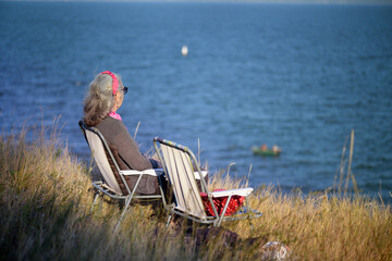 An elderly woman sits on a chair near a lake, a sunny day, autumn time.
