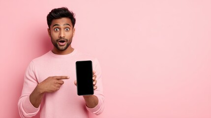 Surprised Indian man holding a smartphone and pointing at the empty screen, astonished expression. Concept of advertising a new mobile app or best deal, isolated on pink background