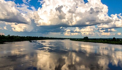 Obraz premium A serene river reflects puffy white clouds against a blue sky. Lush green banks line the water's edge. The light catches the water