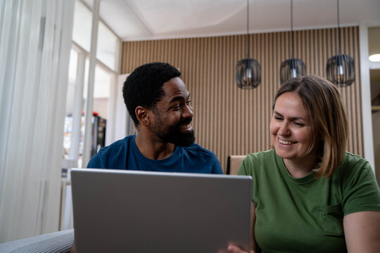 Happy diverse couple smiling and looking at laptop in bright living room during daytime, enjoying love and positive mood.	