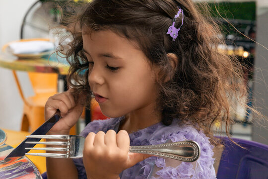 Little toddler girl sitting at the table in summer cafe and holding spoon and fork