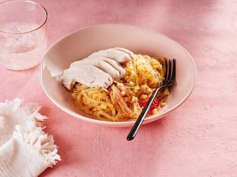 Rice noodles are beautifully plated with healthy slices of turkey. A refreshing glass of water complements the meal, all arranged on a pink surface for a vibrant look