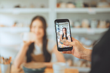 Person taking photo of smiling Asian woman holding handmade ceramic cup in pottery studio using smartphone, social media content creation for small business, lifestyle and creativity concept.