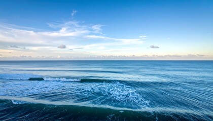 A serene seascape captures rolling ocean waves meeting the horizon under a clear, bright blue sky with scattered wispy clouds. Peaceful atmosphere