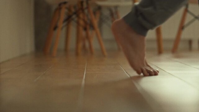 A close-up of a man's feet. Bare feet stepping on wood-patterned tiles in a home kitchen in cozy lighting. A man walks barefoot across the floor of a house. Man's footsteps
