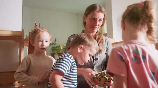 Instructor holds tortoise while curious children gather around observing details of shell and behavior, learning through hands-on interaction in bright classroom setting