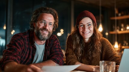 Two cheerful individuals smile for the camera in a cozy space filled with warm lights, showcasing their collaborative spirit and friendship while working on a project together.