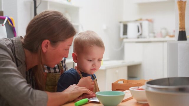 Woman teaching toddlers how to use chopper while guiding hand of boy seated at table, child looking with focus and curiosity, bowls and fruits placed around