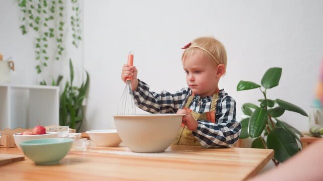 Little one stirs flour with whisk in large bowl during baking preparation, wearing apron and focusing intently, surrounded by kitchen bowls and utensils while learning cooking basics