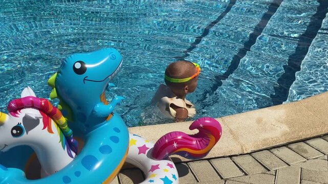 A joyful toddler, wearing a swimming vest and goggles, splashes happily in a bright blue swimming pool alongside colorful inflatable toys. It's a sunny day perfect for a fun water adventure.