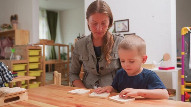 kindergarten teacher guides boy through number recognition with wooden placards, pointing to digit while child traces surface, focused preschool lesson, early math practice builds motor skills