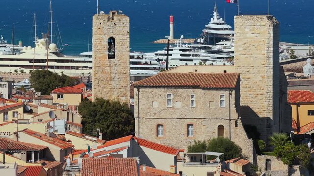 Aerial drone view of terracotta rooftops in Antibes Old Town with the medieval bell tower of Chateau Grimaldi, luxury yachts docked in the marina behind