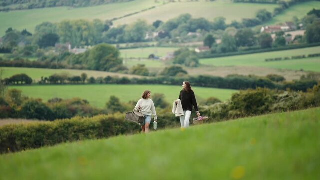 Two women walking in a field with a green background. One of them is holding a surfboard