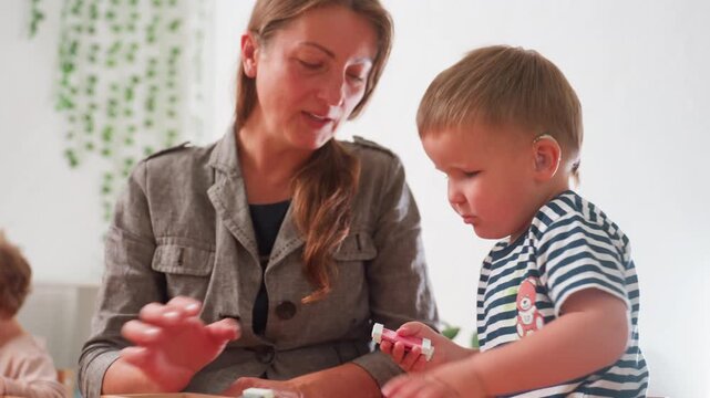 Teacher assisting special child wearing striped shirt as he focuses on small object during classroom activity, supportive presence beside child, warm environment