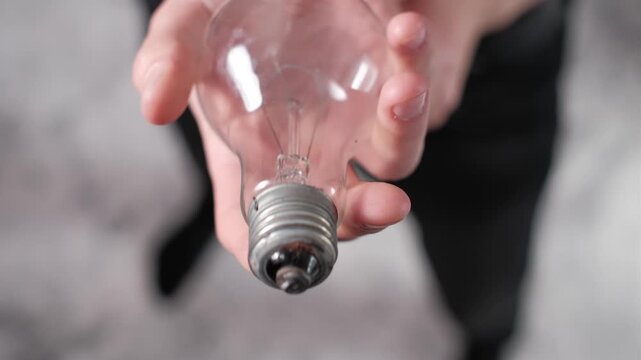 Point-of-view shot of a hand holding a classic incandescent lightbulb and moving it towards the camera, offering or presenting the clear glass bulb with visible filament. Focus idea.