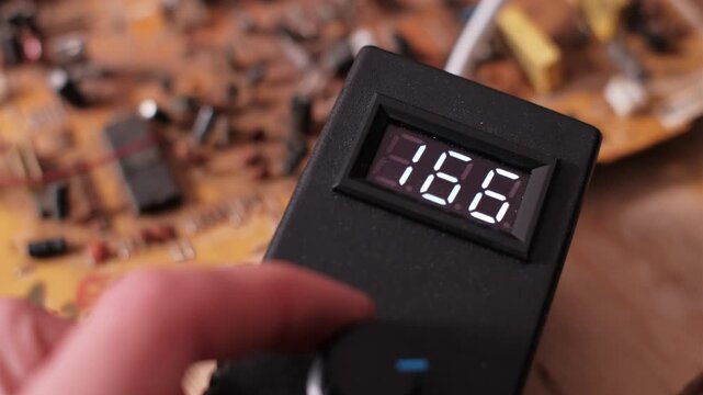 Hand turns a knob on an electronic device, causing the digital display reading to decrease rapidly. Close-up shows voltage change, displaying electrical value numbers in a workshop setting.