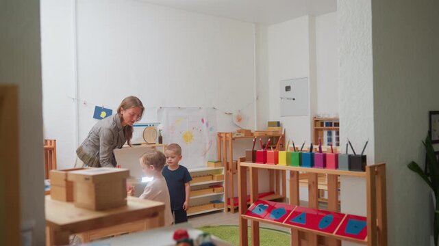 montessori classroom scene with woman teacher setting chair for kids, guiding two children to fetch materials, bright organized room with shelves, art on wall, colorful learning tools