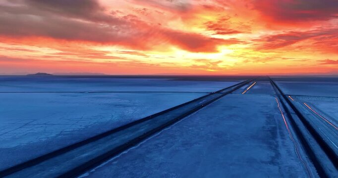 Straight parallel highways crossing the blue landscape. Cars ride by the roads in Bonneville Salt Flats, Utah, USA. Spectacular orange cloudy sky at backdrop.