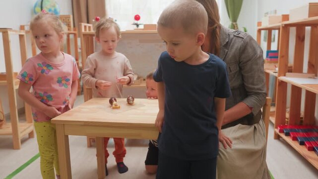 boy in blue seeks attention from teacher during classroom lesson while educator sits guiding group, child looks upset, toddler dynamics, crowding near desk, learning environment