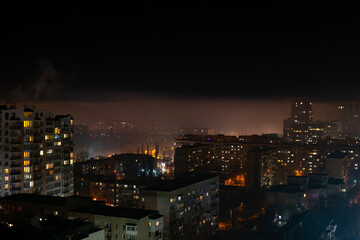 Night cityscape with glowing residential buildings beneath a surreal fog layer. Above the mist, a modern skyline emerges, creating a dramatic contrast and dreamlike atmosphere.