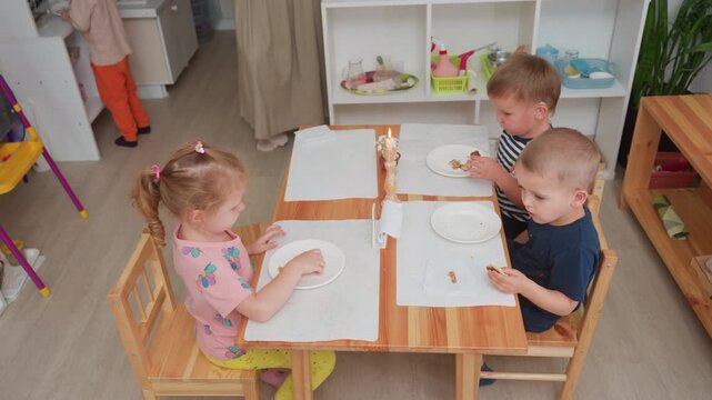 Boy stands to return empty plate after finishing food while other children remain seated eating at wooden table with candle and placemats, classroom environment visible with open shelves and utensils