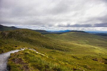 Fototapeta premium Scenic mountainous landscape under dramatic cloudy skies, with a grassy path winding through the rolling hills.