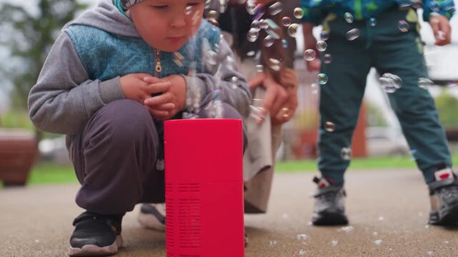 Curious boy squats closely near soap bubble machine watching bubbles rise with fascination as another child approaches from behind, expressing wonder and early curiosity during outdoor park activity
