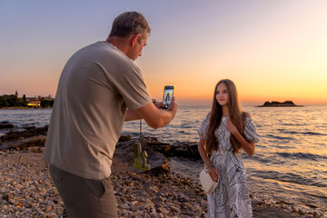 Father taking photo of daughter at sunset beach. The man captures a picture of his daughter posing on a rocky beach during a colorful sunset, highlighting family moments and vacation vibes.