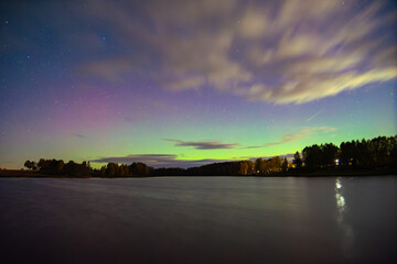 Colorful aurora borealis lights shimmer above a calm lake under a starry sky with clouds and forest reflections.